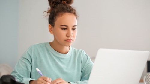 A woman studying with a laptop at home writing notes in a blue top